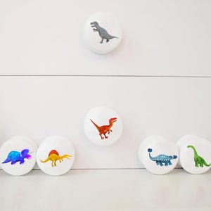 Może przedstawiać: Białe gałki do szafek z kolorowymi ilustracjami dinozaur&oacute;w. Gałki przedstawiają r&oacute;żne wzory dinozaur&oacute;w w odcieniach niebieskiego, zielonego, pomarańczowego i szarego. Te dekoracyjne gałki są idealne do pokoju dziecięcego lub przestrzeni z motywem dinozaur&oacute;w.