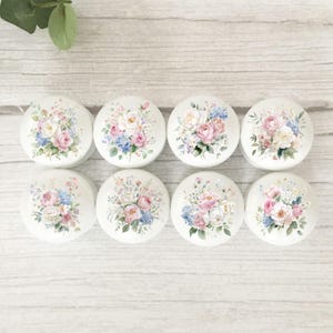 May include: Eight white, round cabinet knobs with floral designs. Each knob features a bouquet of pink, blue, and white flowers. The knobs are arranged on a light wood-grain surface.