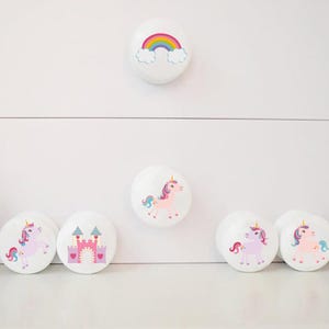 May include: White cabinet knobs with colourful designs. One knob features a rainbow, while others display unicorns and a pink castle. These decorative knobs add a whimsical touch to furniture. The knobs are round and have a smooth finish.