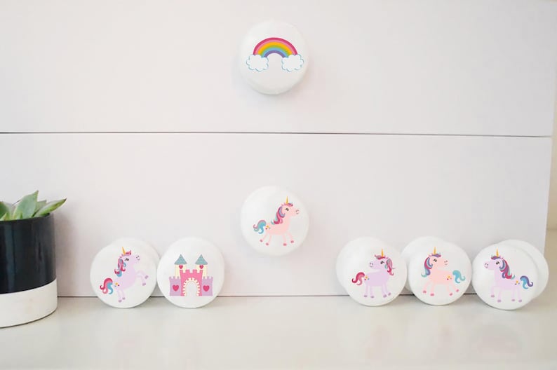 May include: White cabinet knobs with colourful designs. One knob features a rainbow, while others display unicorns and a pink castle. These decorative knobs add a whimsical touch to furniture. The knobs are round and have a smooth finish.