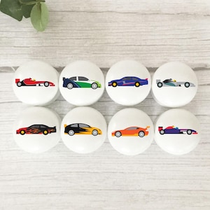May include: Eight white cabinet knobs with race car illustrations. Each knob displays a different car design in colours such as red, black, blue, green, orange, and purple. The knobs are round and have a smooth finish.