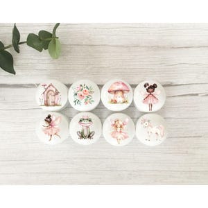 May include: Eight round white ceramic knobs with pink and white fairy themed designs. The designs include a fairy, a frog, a mushroom, a flower, a unicorn, a house, and two fairies.
