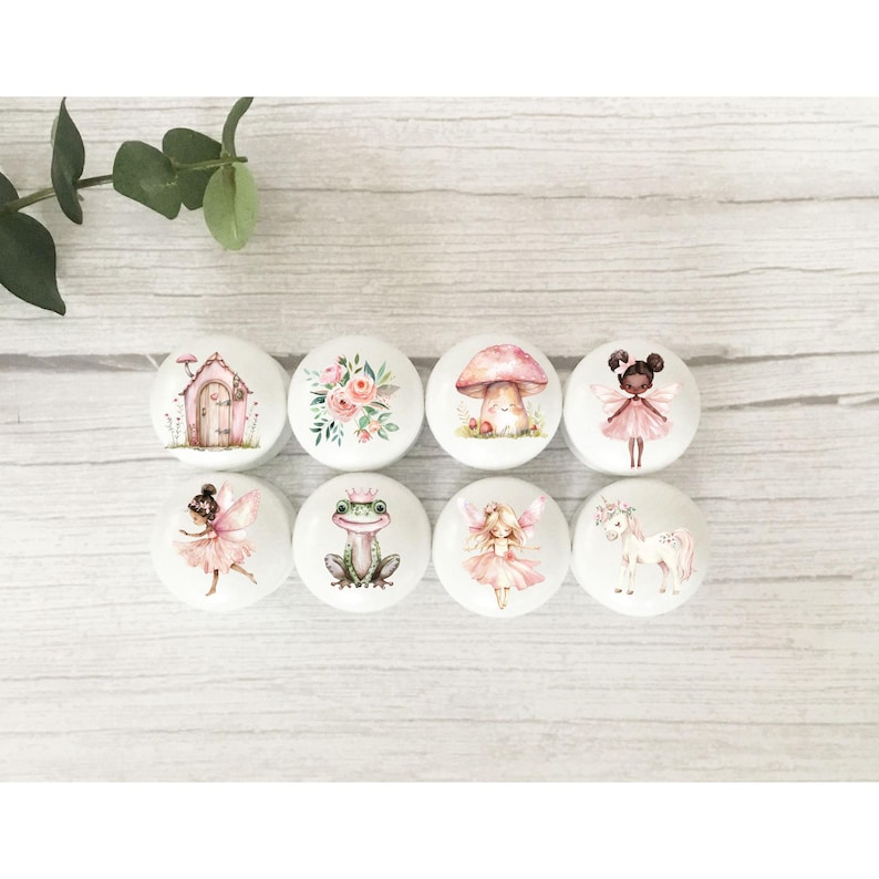 May include: Eight round white ceramic knobs with pink and white fairy themed designs. The designs include a fairy, a frog, a mushroom, a flower, a unicorn, a house, and two fairies.