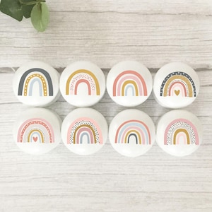 May include: Eight white ceramic cabinet knobs with rainbow designs in pastel colours. Each knob features a different rainbow pattern, including dots and hearts. The knobs are arranged on a light wood surface.