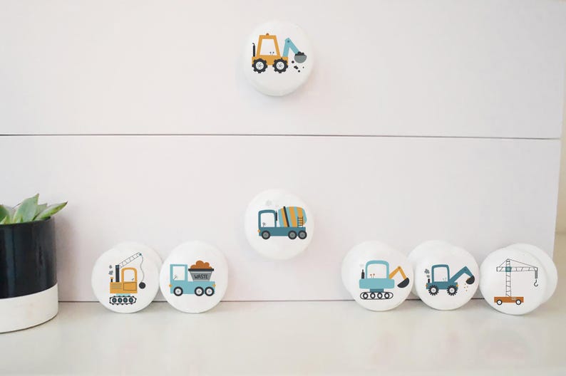 May include: Set of 7 white round drawer knobs with colourful construction vehicle designs. The knobs feature a cement mixer, a dumper truck, a crane, a bulldozer, and two excavators.
