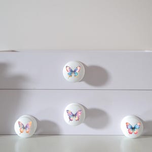 Four white round solid beech furniture knobs each with a different rainbow butterfly illustration. The illustrations are a water colour style with a variety of rainbow gradients. The drawer handles have a glossy finish and are shown on a chest of drawers with succulent plants for decoration.