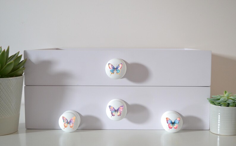 Four white round solid beech furniture knobs each with a different rainbow butterfly illustration. The illustrations are a water colour style with a variety of rainbow gradients. The drawer handles have a glossy finish and are shown on a chest of drawers with succulent plants for decoration.