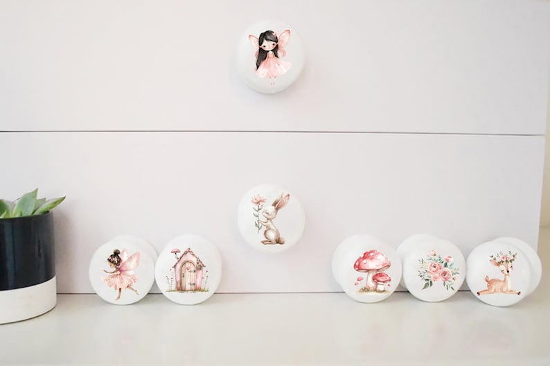 May include: Set of 7 white ceramic drawer knobs with various woodland creature and nature designs. The designs include a fairy, a rabbit, a mushroom, a birdhouse, a floral bouquet, and a deer.
