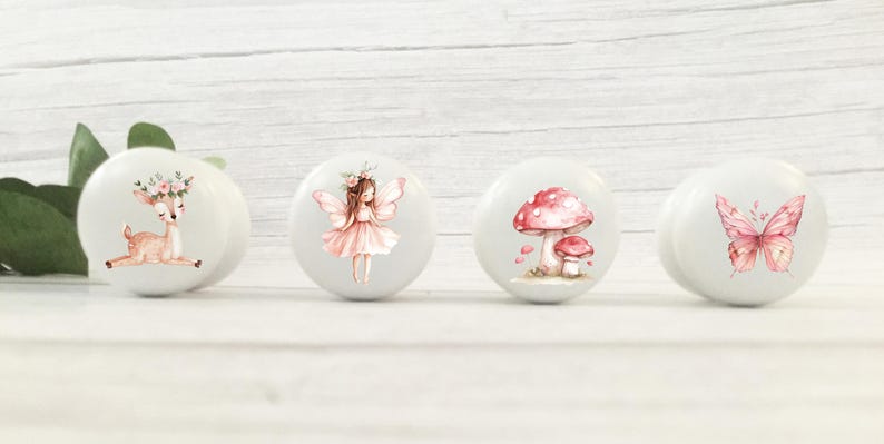 May include: Four white ceramic knobs with pink and brown woodland creature designs. The knobs feature a deer with a floral crown, a fairy with wings, a mushroom, and a butterfly.