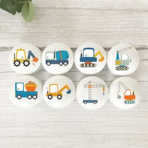 May include: Eight white ceramic cabinet knobs featuring colourful illustrations of construction vehicles. Designs include a yellow excavator, a blue cement truck, and a green crane. Ideal for a child's room or construction-themed space.