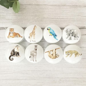 May include: A set of eight round white cabinet knobs, each featuring a different watercolour animal illustration: a tiger, giraffe, blue and yellow macaw, elephant, monkey, zebra, sloth hanging from a branch, and a leopard, arranged in two rows.