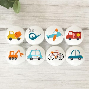 May include: Eight white ceramic cabinet knobs featuring vehicle illustrations. Designs include a yellow cement mixer, a teal helicopter, a blue airplane, a red fire engine, an orange excavator, a blue police car, a red bicycle, and a teal car.