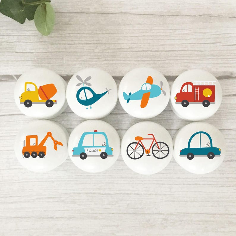 White solid Beech round furniture knobs with bright colourful transport illustrations. The illustrations include a digger, helicopter, aeroplane, fire engine, police car, taxi, bycycle, rocket, cement mixer, cars, road signs, ambulance and hot air balloon. The furniture knobs have a glossy finish.