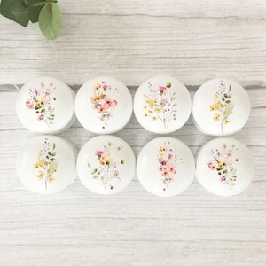 May include: Eight white cabinet knobs with floral designs in pink, yellow, and green. The knobs are round and feature watercolour-style flower arrangements. The background is a light wood surface.
