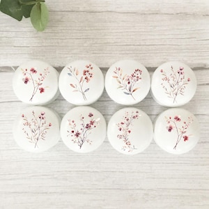 May include: Eight white, round cabinet knobs with floral designs. Each knob features a different watercolour-style botanical illustration in shades of pink, burgundy, and muted green. The knobs are arranged on a light wood-grain surface.