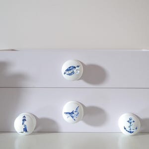 Four white round solid beech furniture knobs each with a different sea life themed illustration. The illustrations include a turtle, shark lighthouse and an anchor. The dresser knobs have a glossy finish and are shown on a chest of drawers with succulent plants for decoration.