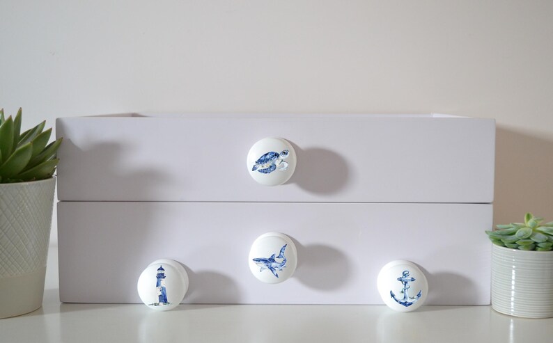 Four white round solid beech furniture knobs each with a different sea life themed illustration. The illustrations include a turtle, shark lighthouse and an anchor. The dresser knobs have a glossy finish and are shown on a chest of drawers with succulent plants for decoration.