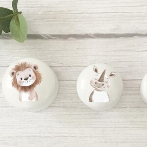 Puede incluir: Tres pomos decorativos redondos blancos con ilustraciones de animales. Un le&oacute;n, un rinoceronte y una jirafa est&aacute;n representados en estilo acuarela. Los pomos est&aacute;n colocados sobre una superficie clara con vetas de madera, con hojas verdes arriba a la izquierda.