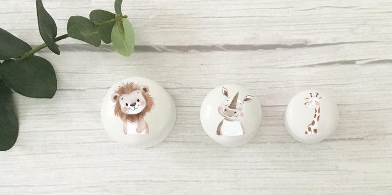 Puede incluir: Tres pomos decorativos redondos blancos con ilustraciones de animales. Un le&oacute;n, un rinoceronte y una jirafa est&aacute;n representados en estilo acuarela. Los pomos est&aacute;n colocados sobre una superficie clara con vetas de madera, con hojas verdes arriba a la izquierda.