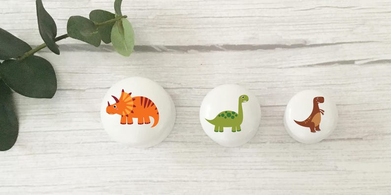 Three white solid Beech furniture knobs with colourful dinosaur designs. The dinosaurs include an orange Triceratops, a brown T Rex and a green Brachiosaurus. The drawer handles are shown in three sizes from largest to smallest and have a smooth glossy finish.
