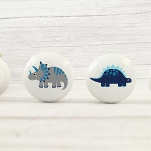 以下が含まれることがあります： 異なる恐竜のデザインが施された、白いセラミック製の引き出しノブが4つ。ノブには、緑色の恐竜、青色のアクセントが付いた灰色の恐竜、青色のアクセントが付いた青色の恐竜、緑色のプテラノドンが描かれています。