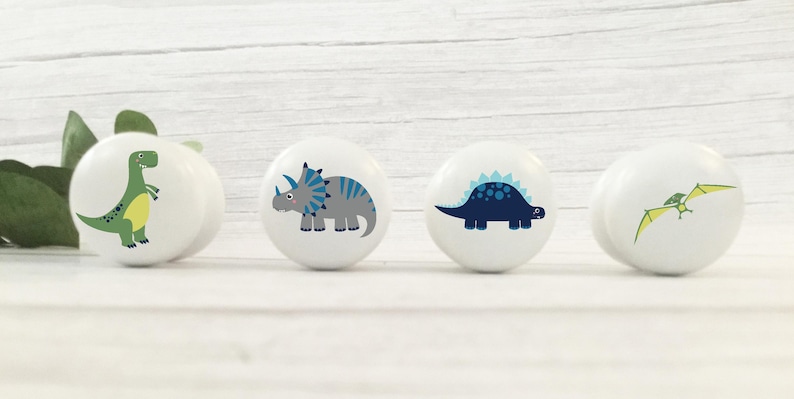 以下が含まれることがあります： 異なる恐竜のデザインが施された、白いセラミック製の引き出しノブが4つ。ノブには、緑色の恐竜、青色のアクセントが付いた灰色の恐竜、青色のアクセントが付いた青色の恐竜、緑色のプテラノドンが描かれています。