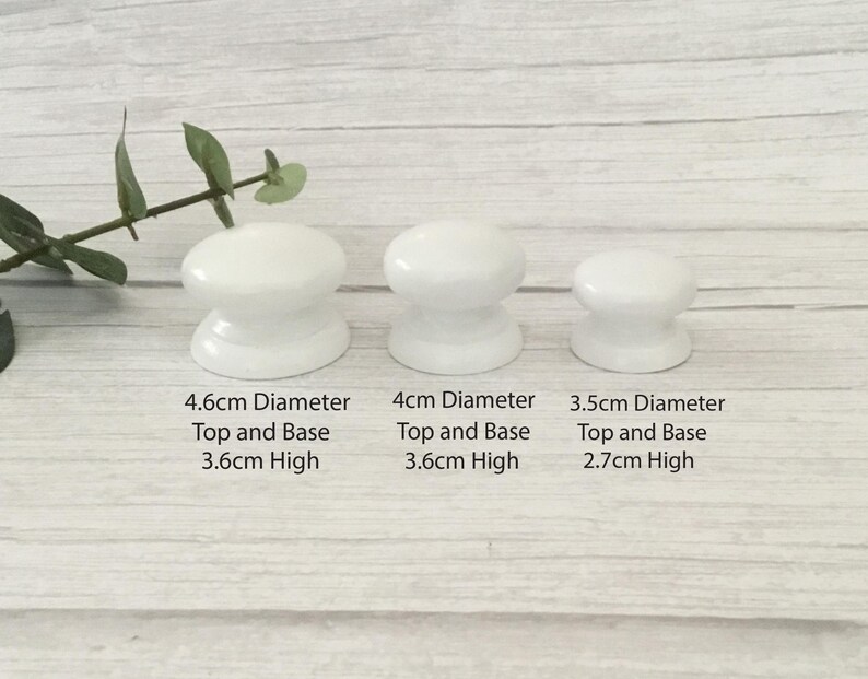 Może przedstawiać: Trzy białe ceramiczne gałki do szafek o r&oacute;żnych rozmiarach. Największa ma średnicę 4,6 cm i wysokość 3,6 cm. Środkowa gałka ma średnicę 4 cm i wysokość 3,6 cm. Najmniejsza ma średnicę 3,5 cm i wysokość 2,7 cm.