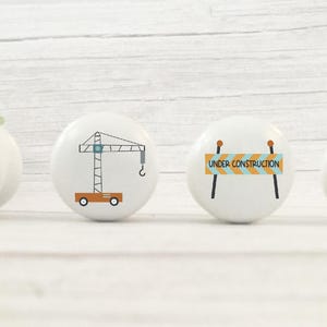 May include: Set of four white ceramic knobs with construction themed illustrations. The knobs feature a yellow crane, a blue excavator, a yellow construction vehicle, and a yellow and blue construction sign that reads "Under Construction".