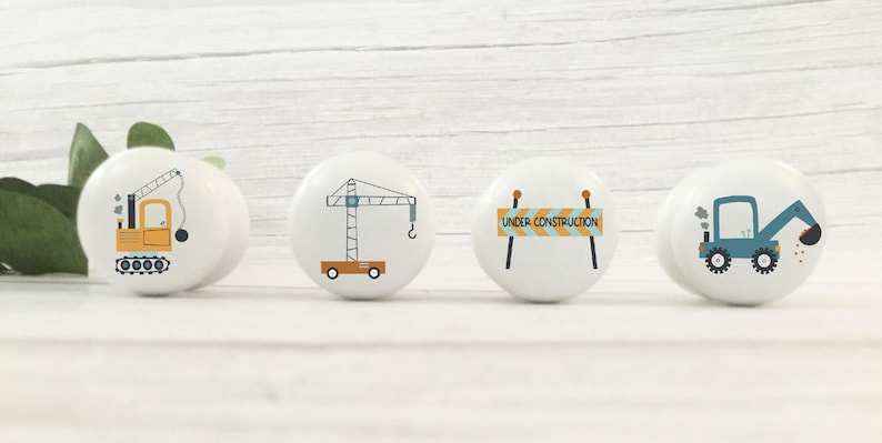 May include: Set of four white ceramic knobs with construction themed illustrations. The knobs feature a yellow crane, a blue excavator, a yellow construction vehicle, and a yellow and blue construction sign that reads "Under Construction".