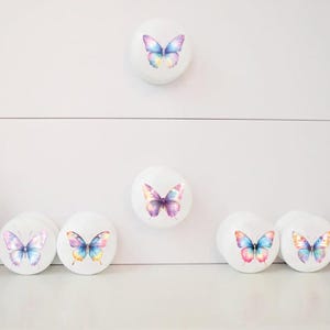 Seven white round solid beech furniture knobs each with a pastel rainbow butterfly design. The designs have slightly different colours but coordinate with each other. The dresser handles have a glossy finish and shown on a chest of drawers with succulent plants for decoration.
