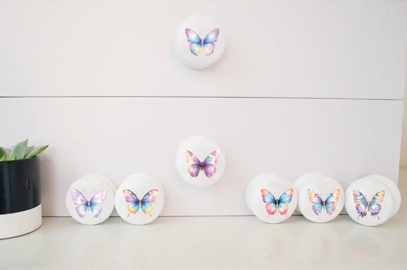 Seven white round solid beech furniture knobs each with a pastel rainbow butterfly design. The designs have slightly different colours but coordinate with each other. The dresser handles have a glossy finish and shown on a chest of drawers with succulent plants for decoration.