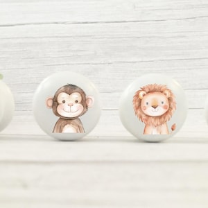 May include: Four white ceramic knobs with animal illustrations. The knobs feature a tiger, monkey, lion, and hippopotamus. The animals are depicted in a cartoon style with smiling faces.