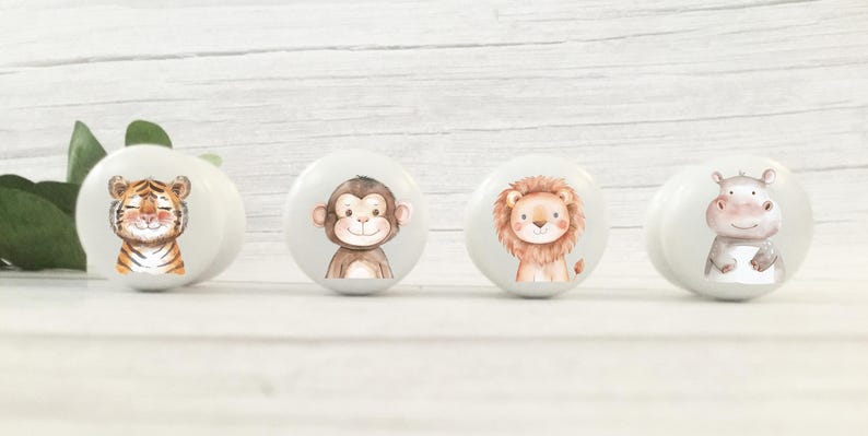 May include: Four white ceramic knobs with animal illustrations. The knobs feature a tiger, monkey, lion, and hippopotamus. The animals are depicted in a cartoon style with smiling faces.