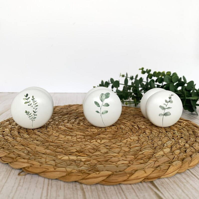 Three round white solid beech furniture knobs are shown each with a different leaf design in different shades of green. They are pictured laying down on a wicker mat with foliage for decoration in the background. The cabinet knobs have a glossy finish.