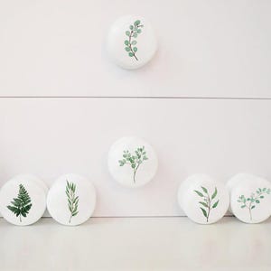 K&ouml;nnte beinhalten: Satz von sieben wei&szlig;en Keramik-Schubladengriffen mit verschiedenen gr&uuml;nen Blattdesigns. Die Griffe sind rund und haben eine glatte, gl&auml;nzende Oberfl&auml;che.