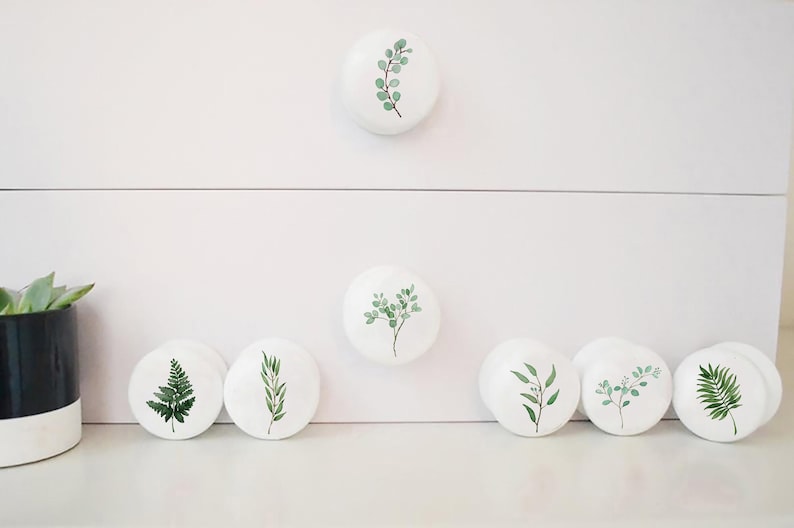 K&ouml;nnte beinhalten: Satz von sieben wei&szlig;en Keramik-Schubladengriffen mit verschiedenen gr&uuml;nen Blattdesigns. Die Griffe sind rund und haben eine glatte, gl&auml;nzende Oberfl&auml;che.