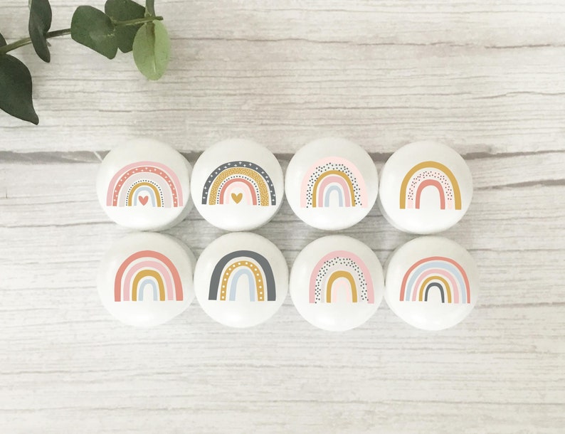 Peut inclure: Huit boutons ronds en c&eacute;ramique blanche avec des motifs arc-en-ciel. Chaque bouton pr&eacute;sente une combinaison de couleurs diff&eacute;rente de rose, bleu, jaune et gris avec des pois noirs. Certains boutons ont un petit c&oelig;ur dor&eacute; au centre.