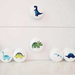 以下が含まれることがあります： カラフルな恐竜のイラストが描かれた白いセラミック製の引き出しノブ6個セット。ノブには、青いT-Rex、緑のトリケラトプス、青いステゴサウルス、黄色のステゴサウルス、青いプテラノドン、緑のティラノサウルス・レックスなど、さまざまな恐竜が描かれています。
