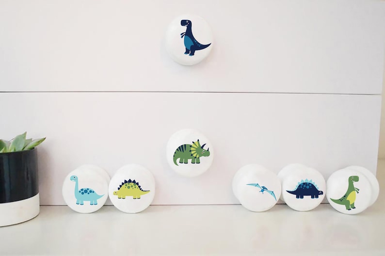 以下が含まれることがあります： カラフルな恐竜のイラストが描かれた白いセラミック製の引き出しノブ6個セット。ノブには、青いT-Rex、緑のトリケラトプス、青いステゴサウルス、黄色のステゴサウルス、青いプテラノドン、緑のティラノサウルス・レックスなど、さまざまな恐竜が描かれています。