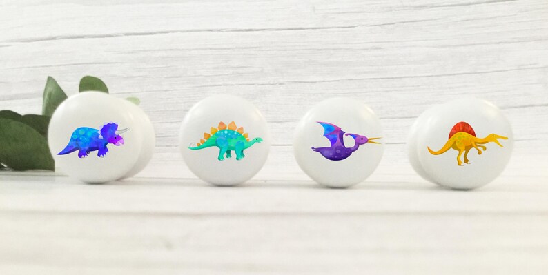 Może przedstawiać: Cztery białe gałki do szafek z kolorowymi ilustracjami dinozaur&oacute;w. Dinozaury to niebiesko-fioletowy triceratops, zielono-niebieski stegozaur, fioletowy pterodaktyl i ż&oacute;łty spinozaur.