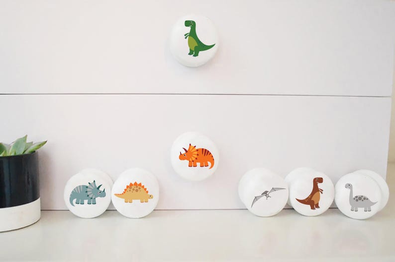 White cabinet knobs featuring dinosaur illustrations in various colours. Designs include a green T-Rex, orange Triceratops, and other prehistoric creatures. Ideal for a child's bedroom or nursery.