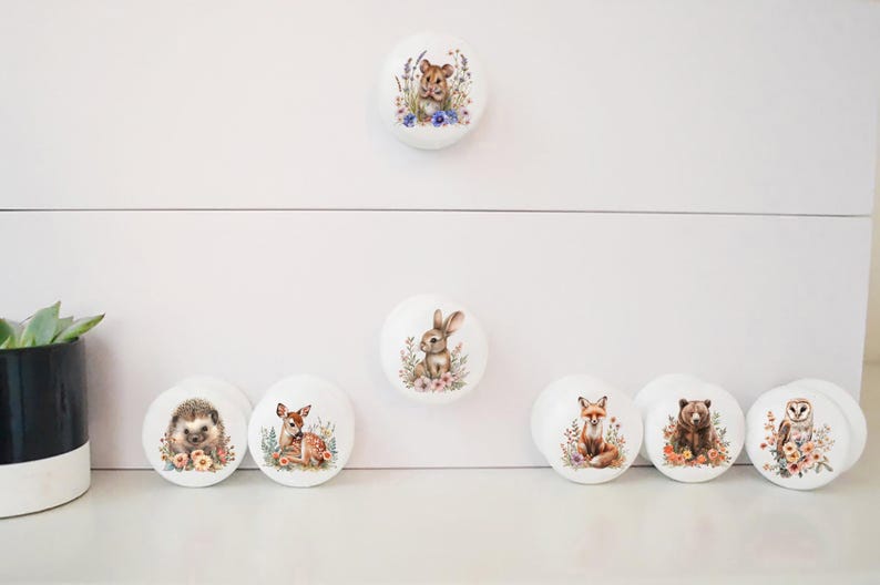 May include: White ceramic cabinet knobs featuring woodland animal illustrations. Designs include a lion, rabbit, hedgehog, deer, fox, bear, and owl, each surrounded by floral accents. Ideal for a nursery or children's room.