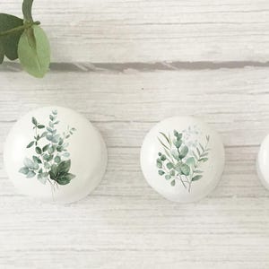 K&ouml;nnte beinhalten: Drei wei&szlig;e, runde dekorative Steine mit Aquarell-Eukalyptusblatt-Designs. Die Bl&auml;tter sind in verschiedenen Gr&uuml;nt&ouml;nen gehalten. Die Steine sind auf einer wei&szlig;en Holzoberfl&auml;che angeordnet, mit einem Eukalyptuszweig im Hintergrund.