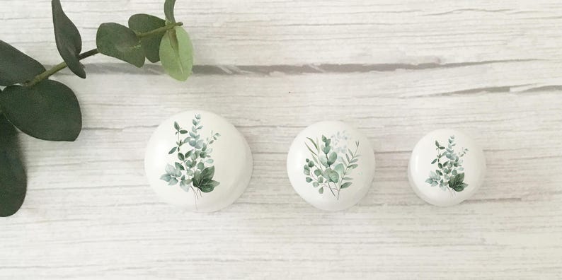 K&ouml;nnte beinhalten: Drei wei&szlig;e, runde dekorative Steine mit Aquarell-Eukalyptusblatt-Designs. Die Bl&auml;tter sind in verschiedenen Gr&uuml;nt&ouml;nen gehalten. Die Steine sind auf einer wei&szlig;en Holzoberfl&auml;che angeordnet, mit einem Eukalyptuszweig im Hintergrund.