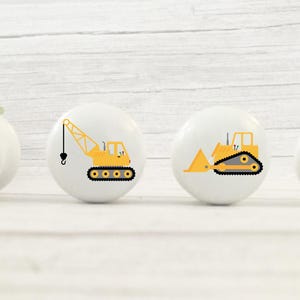 Op de afbeelding: Vier witte keramische knoppen met gele bouwvoertuigontwerpen. De knoppen tonen een betonmixer, een kraan, een bulldozer en een kiepwagen.
