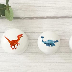 Może przedstawiać: Trzy białe gałki do szafek z ilustracjami dinozaur&oacute;w. Jedna gałka przedstawia pomarańczowego raptora, druga niebieskiego ankylozaura, a ostatnia szarego T-Rexa. Gałki znajdują się na białej drewnianej powierzchni.