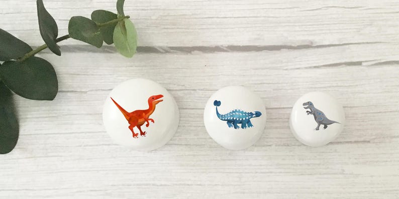 Może przedstawiać: Trzy białe gałki do szafek z ilustracjami dinozaur&oacute;w. Jedna gałka przedstawia pomarańczowego raptora, druga niebieskiego ankylozaura, a ostatnia szarego T-Rexa. Gałki znajdują się na białej drewnianej powierzchni.