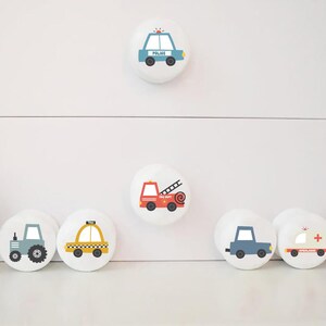 May include: White cabinet knobs with various vehicle designs, such as a police car, fire engine, tractor, taxi, ambulance, and car. The knobs are round and feature simple, cartoon-style illustrations.