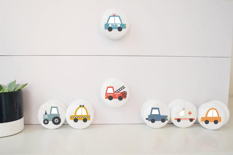 May include: White cabinet knobs with various vehicle designs, such as a police car, fire engine, tractor, taxi, ambulance, and car. The knobs are round and feature simple, cartoon-style illustrations.