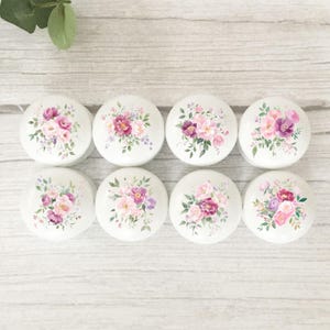 May include: Eight white cabinet knobs with floral designs. Each knob features a different arrangement of pink, purple, and white flowers with green leaves. The knobs are arranged on a light wood surface.